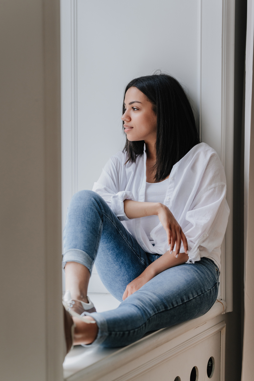 Sad Brazilian young woman sitting on windowsill in white shirt and blue jeans frustrated after divorce, thinking about future holding phone at home, sunny day. Pretty African American girl in troubles